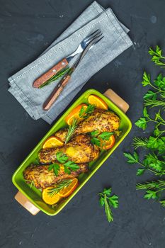 Baked chicken drumstick in a green dish with orange and rosemary, black stone concrete surface, top view, copy space.