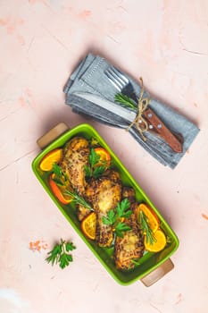 Baked chicken drumstick in a green dish with orange and rosemary, light pink living coral stone concrete surface, top view, copy space.