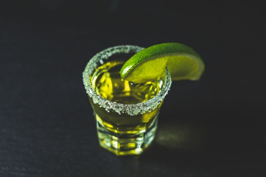 Mexican Gold Tequila shot  with lime and salt on black stone table surface, selective focus, shallow depth of the field, copy space.