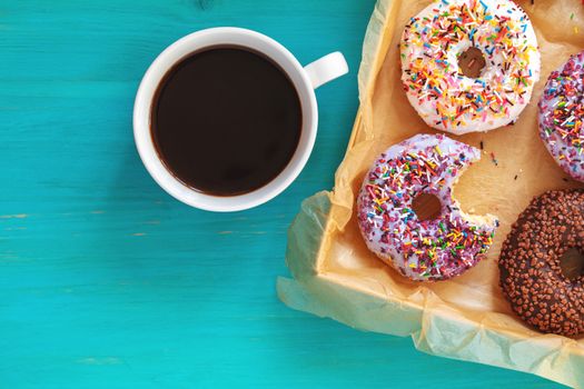 Delicious glazed donuts in box and cup of coffee on turquoise blue surface. Flat lay minimalist food art background. Top view.