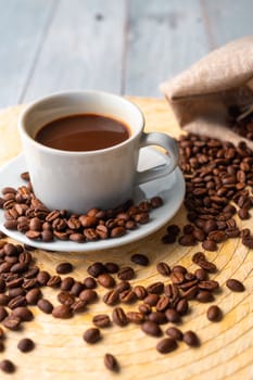 White cup and saucer with coffee and surrounded by roasted coffee beans on a wooden table and a bag