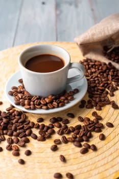 White cup and saucer with coffee and surrounded by roasted coffee beans on a wooden table and a bag