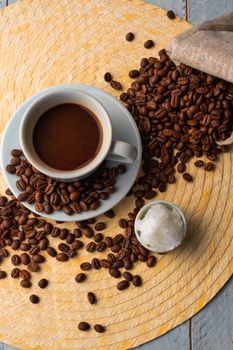 White cup and saucer with coffee and surrounded by roasted coffee beans on a wooden table and a bag next to a small cup of coconut oil