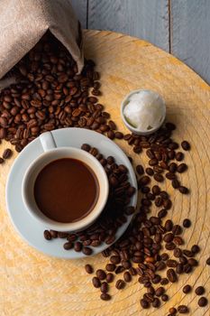 White cup and saucer with coffee and surrounded by roasted coffee beans on a wooden table and a bag next to a small cup of coconut oil