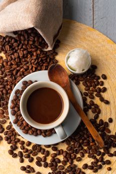 White cup and saucer with coffee and surrounded by roasted coffee beans on a wooden table and a bag next to a small cup of coconut oil and a wooden spoon