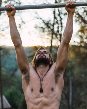 Young bearded man doing pull ups in the park with beautiful sunset on the mountain