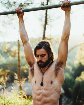 Young bearded man doing pull ups in the park with beautiful sunset on the mountain