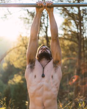 Young bearded man doing pull ups in the park with beautiful sunset on the mountain