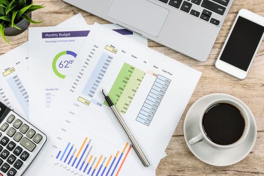 Overhead view of a modern workplace at the office: printouts of charts and financial data next to a laptop, smartphone and calculator on the desk.