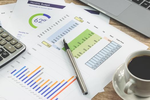 Modern workplace at the office: printouts of charts and financial data next to a laptop, cup of coffee and calculator on the desk (reduced tone)