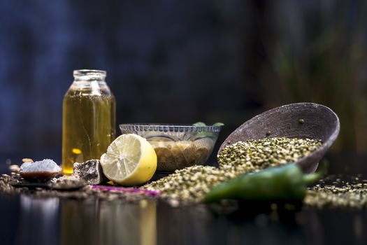 Close up of mung dal soup in glass bowl along with entire constituents with it which are mung bean,green gram, asafoetida, cumin, green chili, black peppercorns, lemon juice and salt on black surface.