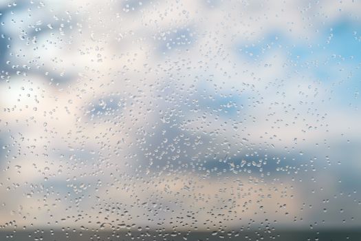 Raindrops on a window glass against blue sky with clouds.