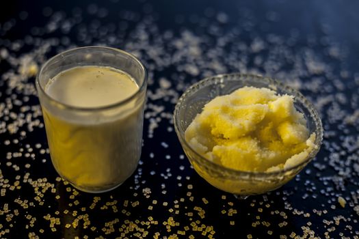 Close up of glass bowl of pure milk well mixed with hot milk in it on black wooden glossy surface along with raw ghee clarified butter and some sugar crystals spread on the surface. Horizontal shot.;