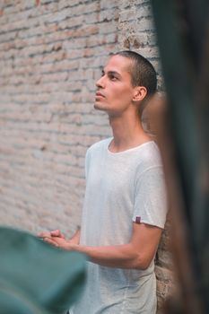 Exotic caucasian young man with a white shirt looking to the side posing in front of a brick wall and behind a big tropical plant