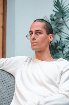 Exotic caucasian young man with round transparent sunglasses looking to the camera while sitting on a sofa in front of a tropical plant