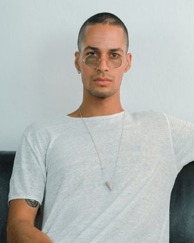 Exotic caucasian young man with round transparent sunglasses looking to the camera while sitting on a sofa with white background