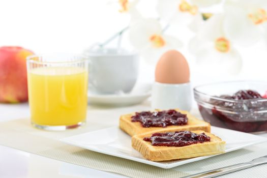 Crispy toast with raspberry jam on a plate and a soft-boiled egg next to orange juice, coffee and apples on the background of blooming orchid