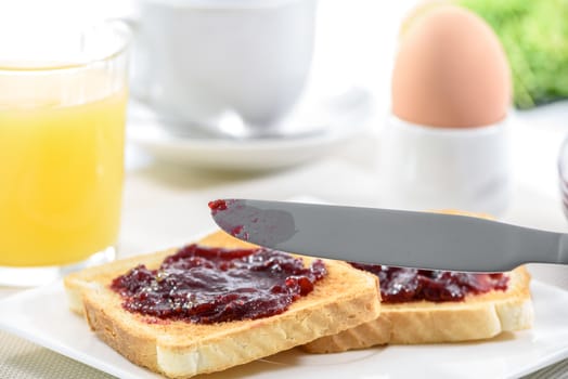 Delicious breakfast with two toast on a plate with jam, a knife on the front and orange juice with egg behind. 
