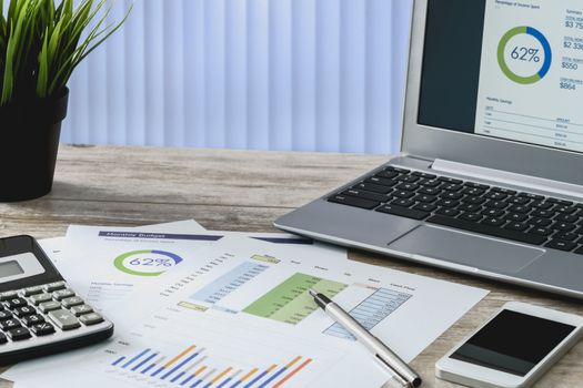 Modern workplace at the office: printouts of charts and financial data next to a laptop, smartphone and calculator on the desk.