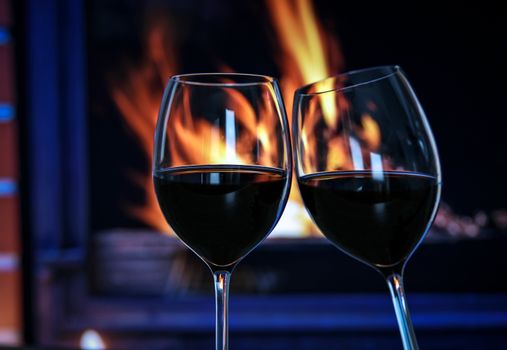 Two glasses of red wine raised in a toast against a fire in the fireplace.