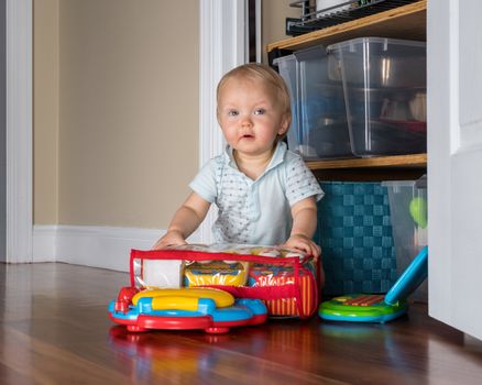 Baby boy sitting on the wooden floor of living room and playing with two toy computers and some toy cars