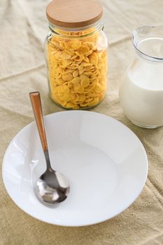 Cornflakes in a jar, next to a transparent jug of milk, an empty plate with a spoon on a plain rough tablecloth. View from above