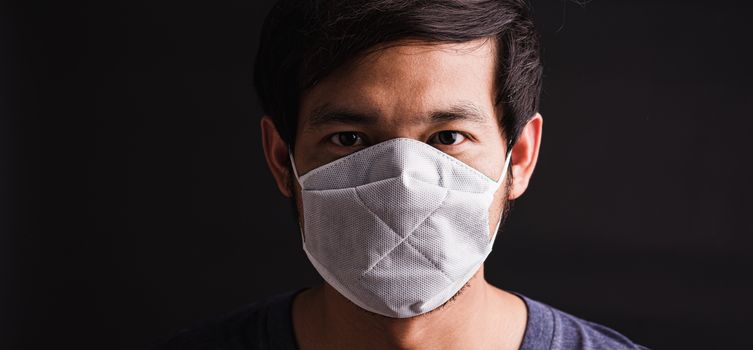 Closeup Asian young man wearing protective face mask with fear in the eye, prevent germs or disease hygiene prevention COVID-19 virus or coronavirus protection concept, dark on black background