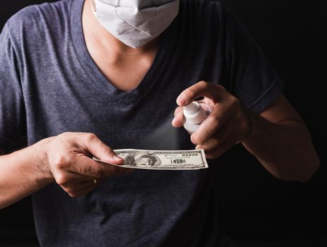 Closeup man wearing protective face mask applying sanitizer spray to 100 dollar money for cleaning, hygiene prevention COVID-19 virus or coronavirus protection concept, dark on black background