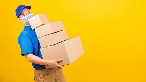 Asian young delivery worker man in blue t-shirt and cap uniform wearing face mask protective lifting stack a lot of boxes, under coronavirus or COVID-19, studio shot isolated yellow background