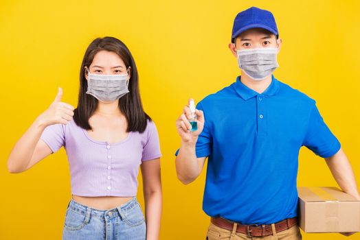 Asian young handsome delivery man hold alcohol disinfectant spray and woman show finger thumb up and both wearing face mask protective  virus, isolated yellow background, outbreak coronavirus COVID-19