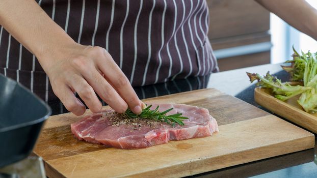 The housewives seasoned the meat pieces with salt, pepper and fresh rosemary before cooking it into a steak. Morning atmosphere in a modern kitchen.