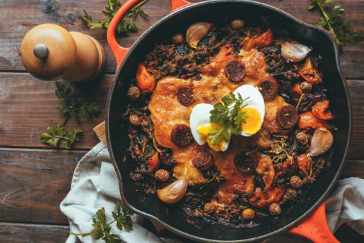 Wrought iron skillet with vegetables, meat, egg, gratin cheese and coriander on a rustic wooden table