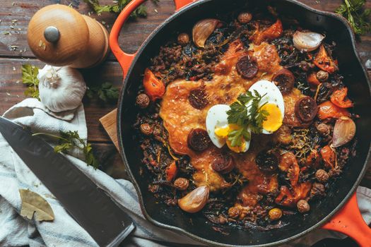 Wrought iron skillet with vegetables, meat, egg, gratin cheese and coriander with a knife on a rustic wooden table