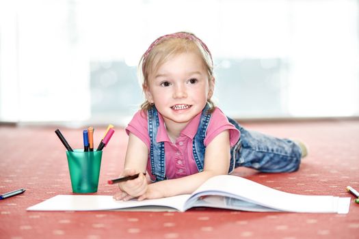 Blond hair girl lying on the carpet and plays with drawing n the children's playroom.