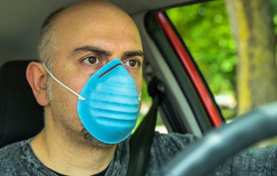 Turin, Piedmont, Italy. April 2020. Coronavirus pandemic: portrait of a Caucasian man driving the car wearing a blue mask to avoid contagion. Selective focus on man and blurred background.