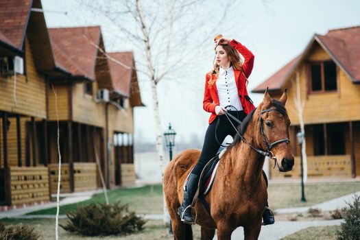 red-haired jockey girl in a red cardigan and black high boots with a horse for a walk