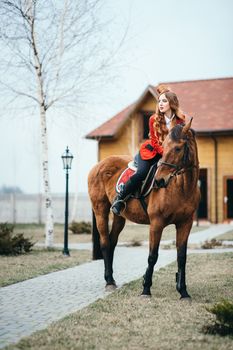 red-haired jockey girl in a red cardigan and black high boots with a horse for a walk