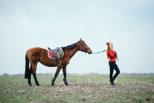 red-haired jockey girl in a red cardigan and black high boots with a horse for a walk