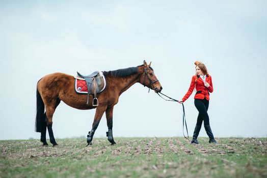 red-haired jockey girl in a red cardigan and black high boots with a horse for a walk