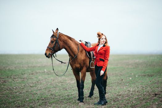 red-haired jockey girl in a red cardigan and black high boots with a horse for a walk