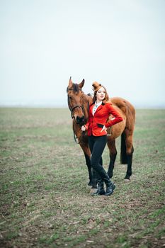 red-haired jockey girl in a red cardigan and black high boots with a horse for a walk