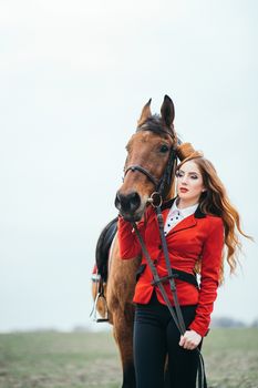 red-haired jockey girl in a red cardigan and black high boots with a horse for a walk