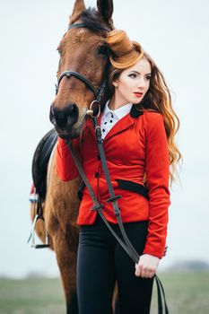 red-haired jockey girl in a red cardigan and black high boots with a horse for a walk