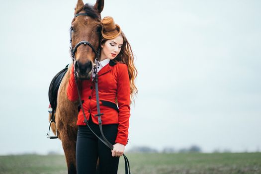 red-haired jockey girl in a red cardigan and black high boots with a horse for a walk