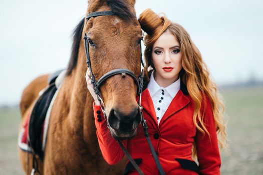 red-haired jockey girl in a red cardigan and black high boots with a horse for a walk