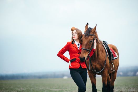 red-haired jockey girl in a red cardigan and black high boots with a horse for a walk