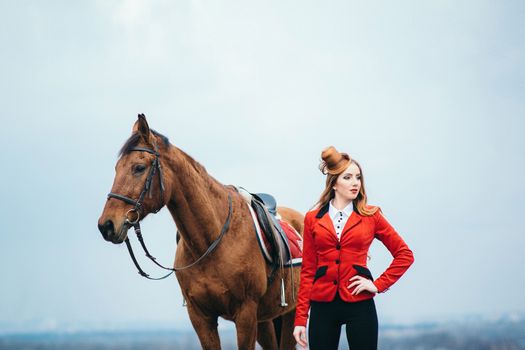 red-haired jockey girl in a red cardigan and black high boots with a horse for a walk