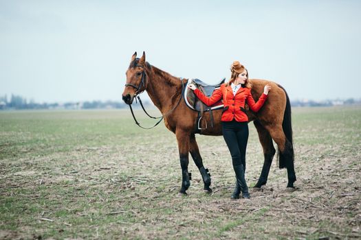 red-haired jockey girl in a red cardigan and black high boots with a horse for a walk