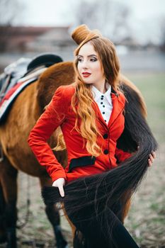 red-haired jockey girl in a red cardigan and black high boots with a horse for a walk