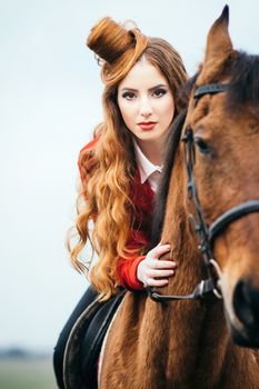 red-haired jockey girl in a red cardigan and black high boots with a horse for a walk